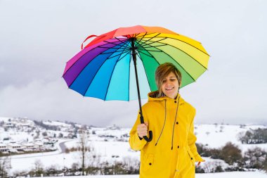 Gökkuşağı renginde bir şemsiye taşıyan mutlu bir kadının portresi bir köyün karında koşuyor ve güzel bir manzarası var ve sarı bir ceket giyiyor çok renkli bir resim yapıyor.
