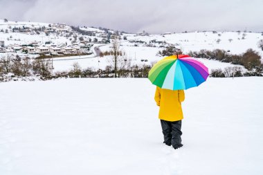 Gökkuşağı renginde bir şemsiye taşıyan bir kadının arka planda güzel bir manzarası olan bir dağda yürüdüğünü ve sarı bir ceket giydiğini ve çok renkli bir resim çizdiğini görüyoruz.