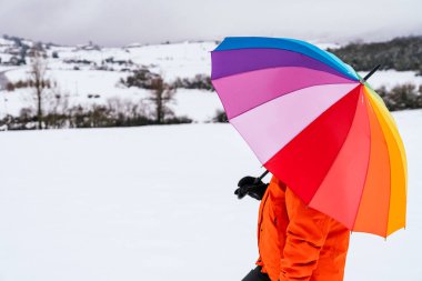 Çok renkli bir şemsiye taşıyan bir kadının portresinin yan görüntüsü güzel bir manzaraya sahip dağdaki gökkuşağı gibi ve çok renkli bir resim yapan turuncu bir ceket giyiyor.
