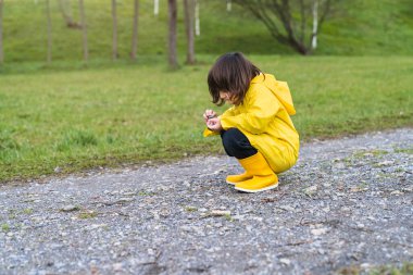 Yağmurluklu ve sarı çizmeli bir çocuk yere çömelmiş taşlarla oynuyor.