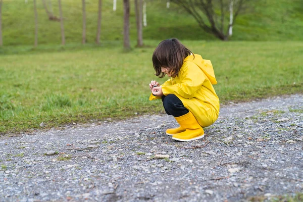 Boy in raincoat and yellow rain boots crouching down playing with some stones on the ground