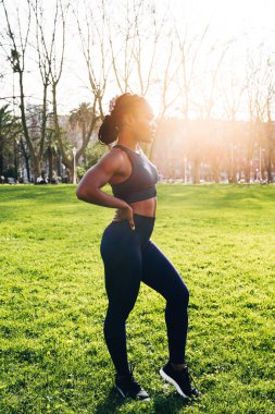 Siyah afro kız gün batımında halka açık bir parkta koşuya çıkmadan önce kol ve sırt esneme hareketleri yapıyor.