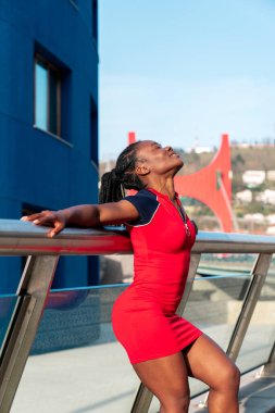 Siyah model Afro kız, güler yüzlü ve eğlenceli bir poz veriyor. Gün batımında kırmızı bir elbise ve beyaz ayakkabılar giyiyor.