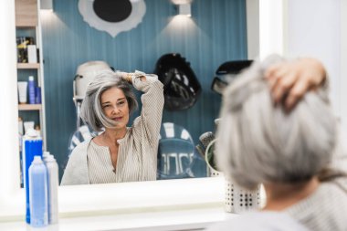 Kıdemli kadın kuaför aynasında yansımaya bakıyor, beyaz saçlarına dokunuyor, doğal güzellik hareketini kucaklıyor, şekillendiriyor.