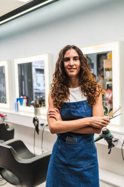 Gülümseyen kadın kuaför önlük takıyor ve elinde makasla modern kuaföründe poz veriyor.