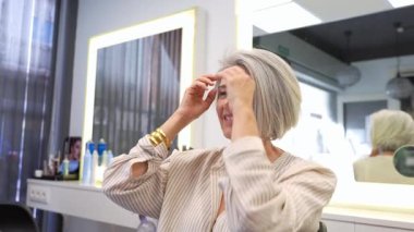 Zarif, olgun bir kadın. Gri saçları var. Gülümsüyor ve yeni saç kesimine dokunuyor.