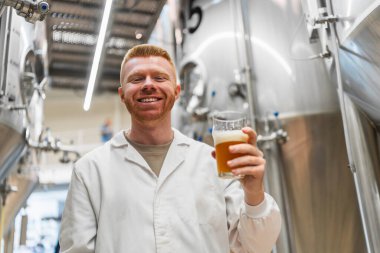 Laboratuar önlüklü gülümseyen adam, elinde kehribar renkli bir bardak bira, çelik tanklarla modern bir bira fabrikasında.