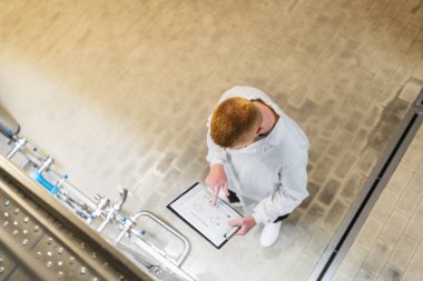 Kalite kontrol için dijital tablet kullanan ve endüstriyel bir tesiste bira üretimini izleyen beyaz önlüklü bir işçi.