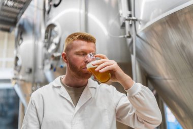 Labaratuvar önlüğünün içinde bira tadımı bira fabrikası üretim alanında bira tatma, paslanmaz çelik tankların arasında lezzet ve kaliteyi denetleme