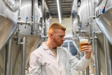 Laboratuvar önlüklü bir adam, bir bardak birayı inceliyor, modern paslanmaz çelik fabrikasında kalite kontrolü sağlıyor.