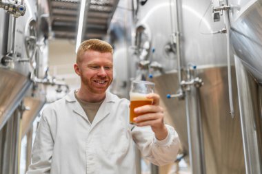 Gülümseyen bira imalatçısı kaliteli bira bardağını inceliyor, paslanmaz çelik tanklarıyla küçük bir bira fabrikasında duruyor.