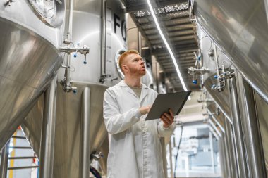 Laboratuvar önlüklü adam büyük mayalama tanklarını kontrol ediyor, modern bir içecek üretim tesisinde kalite kontrolü yapıyor.