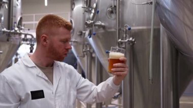 Beyaz önlüklü profesyonel bira üreticisi büyük çelik fermantasyon tanklarıyla modern bir bira fabrikasında el sanatlarının kalitesini inceliyor ve inceliyor.