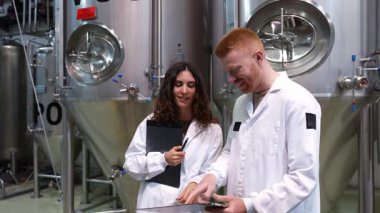 Beyaz önlüklü iki bira fabrikası işçisi üretim verilerini tartışıyor. Erkek ve kadın teknisyenler büyük çelik fermantasyon tanklarının önünde işbirliği yapıyorlar.