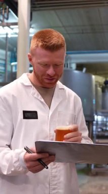 Beyaz önlüklü genç bir bira imalatçısı bira fıçısının rengini, berraklığını ve köpüğünü incelerken bir yandan da bira fabrikasındaki bir panoya not alıyor.