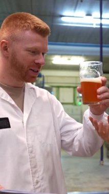 Beyaz önlüklü usta, bir bardak taze birayı tadıyor ve değerlendiriyor. Modern bir fabrikada kalitesini ve tadını kontrol ediyor.