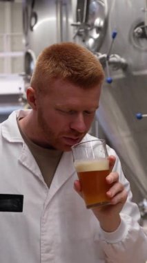 Beyaz önlüklü bira ustası hafif bira kokusu ve tadına bakıyor. Çelik fermantasyon tanklarının yanındaki lezzetin tadını çıkaran adam.