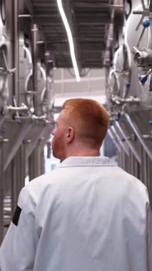 Büyük çelik fermantasyon tankları arasında yürüyen beyaz önlüklü bir erkek bira üreticisi. Endüstriyel bira üretim tesisinin yavaş çekim görüntüleri