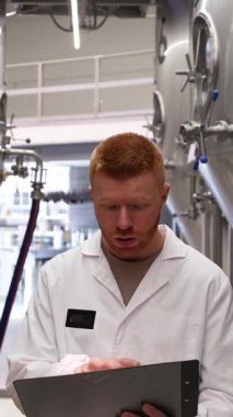 Beyaz laboratuvar önlüklü bir erkek işçi bir fabrikada büyük çelik tankları inceliyor. Elinde bir pano var ve demleme işlemini izliyor.