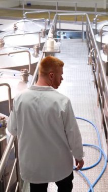 Metal bir platformda yürüyen beyaz önlüklü bir bira ustasının arka görüntüsü. Büyük paslanmaz çelik fermantasyon tanklarını inceliyor.