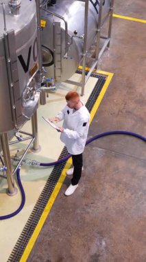 Beyaz ceketli, fermantasyon tanklarını kontrol eden ve yavaş çekimde not alan bir erkek bira fabrikası işçisinin tepeden aşağı bakışı.