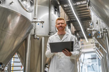Beyaz laboratuvar önlüklü genç adam gülümsüyor, elinde bir pano, bira fabrikasında paslanmaz çelik fermantasyon tanklarının arasında duruyor.
