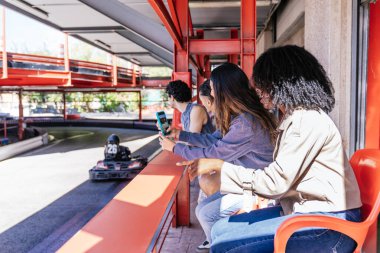 Arkadaşlar go kart pistinde eğleniyor, yarış seyrediyor, akıllı telefonlarla kayıt yapıyor ve birlikte eğleniyorlar.