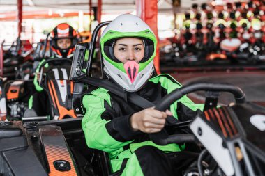 Yarış takımlı ve kasklı genç bir kadın kart sürmeye hazırlanıyor. İlerideki karting pistine odaklanıyor.