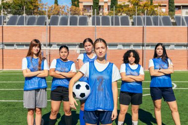 Güneşli bir açık hava spor sahasında takım çalışmasını ve güçlendirmeyi temsil eden çeşitli bayan futbolcular birlikte poz veriyorlar.