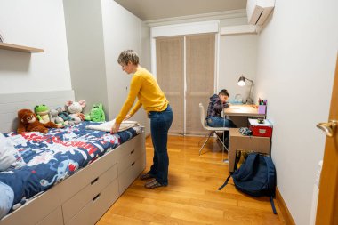 Anne, oğlunun yatağını odasında toplarken çocuk da masa başında ödev yapıyor ve aile rutinini gösteriyor.