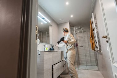 Bir kadın banyoda saç kurutma makinesini fişe takıyor, güne hazırlanıyor ve sabah rutininde saçını şekillendiriyor.