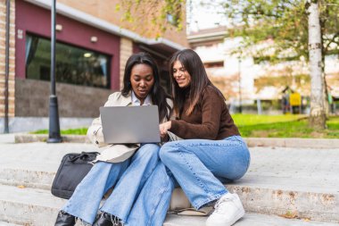 Two young diverse women students sitting on stairs outside, collaborating and discussing while using a laptop computer