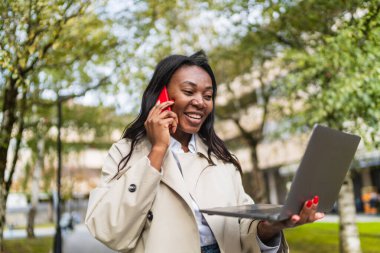 Gülümseyen genç iş kadını birden fazla iş yapıyor, akıllı bir telefonla konuşuyor ve şehir parkında dizüstü bilgisayar tutuyor, esnek bir işi kucaklıyor.