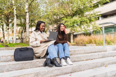 İki farklı kız öğrenci kampüs merdivenlerinde oturuyor, biri dizüstü bilgisayar kullanıyor, diğeri akıllı telefondan konuşuyor, iletişim kuruyor ve çalışıyor.