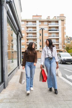 İki farklı arkadaş alışveriş torbalarıyla sokakta yürüyor, birlikte vakit geçirip sohbet ediyorlar.