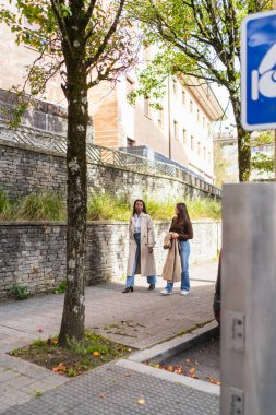 Trençkotlu iki farklı kadın sonbahar şehir kaldırımında yürüyor ve sohbet ediyor, gülümsüyor ve gündüz gezintisinden keyif alıyorlar.