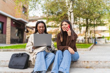 Colleagues or friends sitting outdoors, working on a laptop and talking on a smartphone, embracing remote work and technology