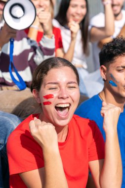 Yüzü boyayla coşkuyla tezahürat eden genç bir kadın. Bir grup farklı arkadaş canlı bir spor stadyumunda kutlama yapıyorlar.