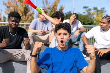 Heyecanlı çoklu etnik gruptan genç erkekler bir spor maçı sırasında takımlarına tezahürat yapıyor, coşku ve hayal kırıklığı gösteriyorlar.