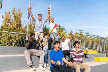 Stadyumda oturan çok ırklı gençler spor maçı izleyip bayrak ve vuvuzela ile kutlama yapıyorlar.