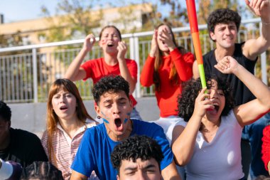 Çeşitli gençler hareketli bir stadyum atmosferinde aktif tepki gösterip bir spor olayını kutluyorlar.