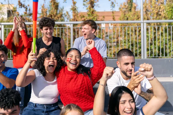 Çok ırklı genç yetişkinler bir stadyumda spor müsabakasını izlerken heyecanla kutlama ve bağırıyorlar.