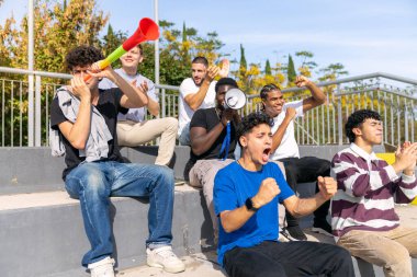 Bir spor müsabakası sırasında takımlarını stadyum tribünlerinde coşkuyla alkışlayan ve kutlayan bir grup genç.
