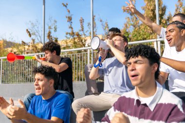 Bir grup erkek taraftar stadyumdan bir spor müsabakasını izliyor, kutlama yapıyor ve birlikte gürültü yapıyorlar.