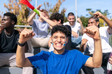 Stadyum tribünlerinde kutlama yapan heyecanlı bir grup arkadaş, takımlarına destek ve coşku gösteriyorlar.