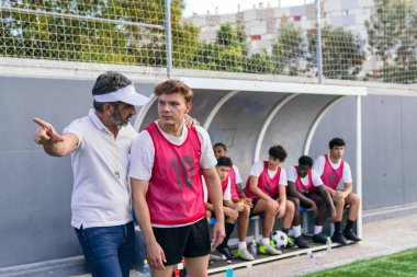 Futbol koçu sahada genç bir oyuncuyu eğitirken takım arkadaşları antrenman sırasında yedek kulübesinde izliyor.