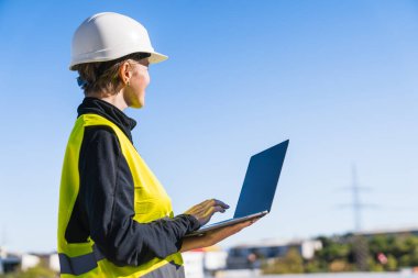 Sert şapka ve güvenlik yeleği giyen kadın mühendis dışarıda dizüstü bilgisayardaki güneş enerjisi üretim verilerini inceliyor.