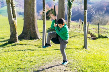 Neşeli çocuk halatla salıncakta aktif olarak oynuyor. Doğal bir oyun alanında, eğlenceli bir an yaşıyor.