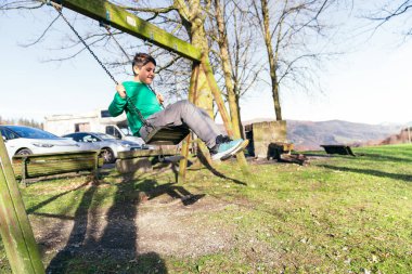 Güneşli bir parkta ahşap bir salıncakta sallanan çocuk, gülüyor ve kaygısız yeşil çimenler, mavi gökyüzü ve çocukluk özgürlüğü sırasında ağaçların arasında.