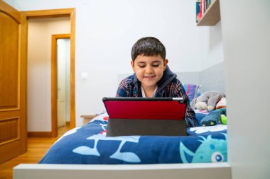 Genç çocuk yatakta yüzüstü yatar, video izler ya da yatak odasındaki dijital tablette oyun oynar.